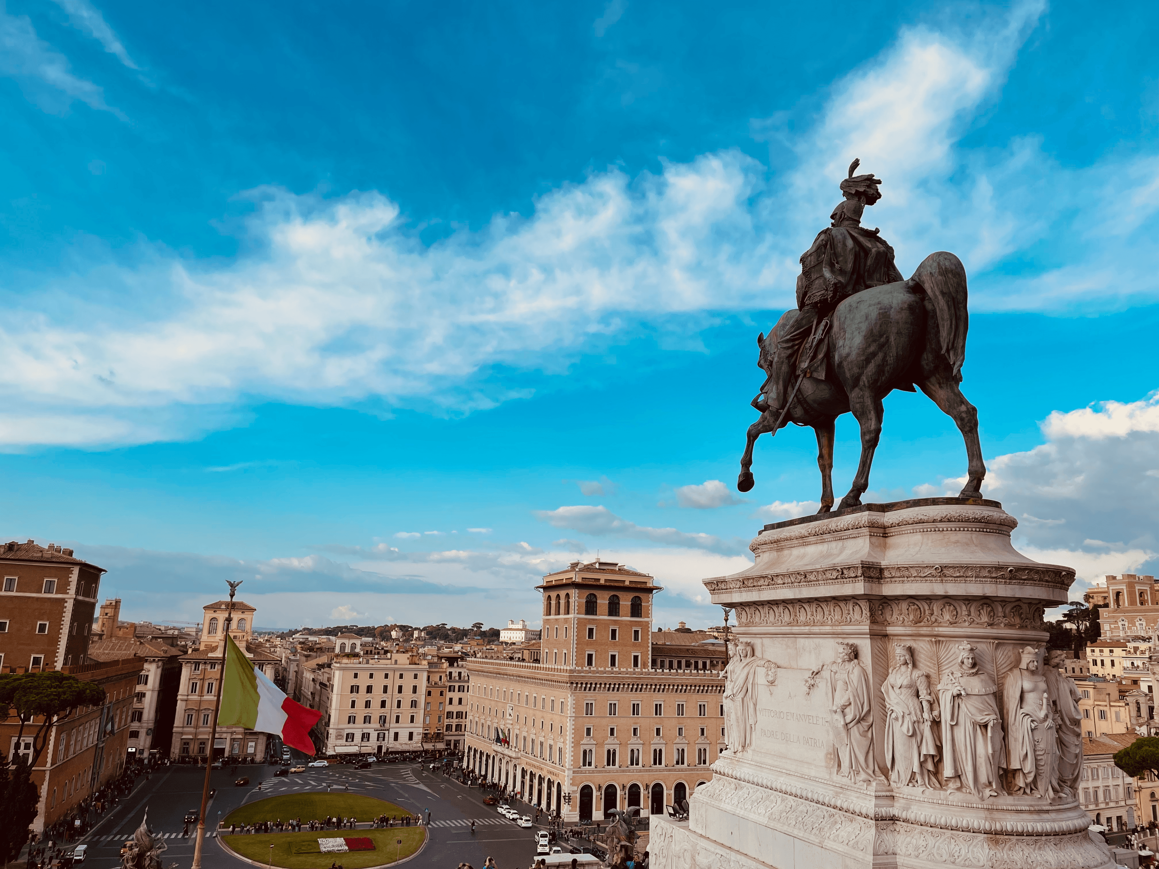 Picture taken from behind the Altar of the Fatherland in Rome, Italy. On the foreground, a statue of the first king of Italy mounted on a horse. In the background, the italian flag and the cityscape of Rome, with old, historical buildings below a blue sky.